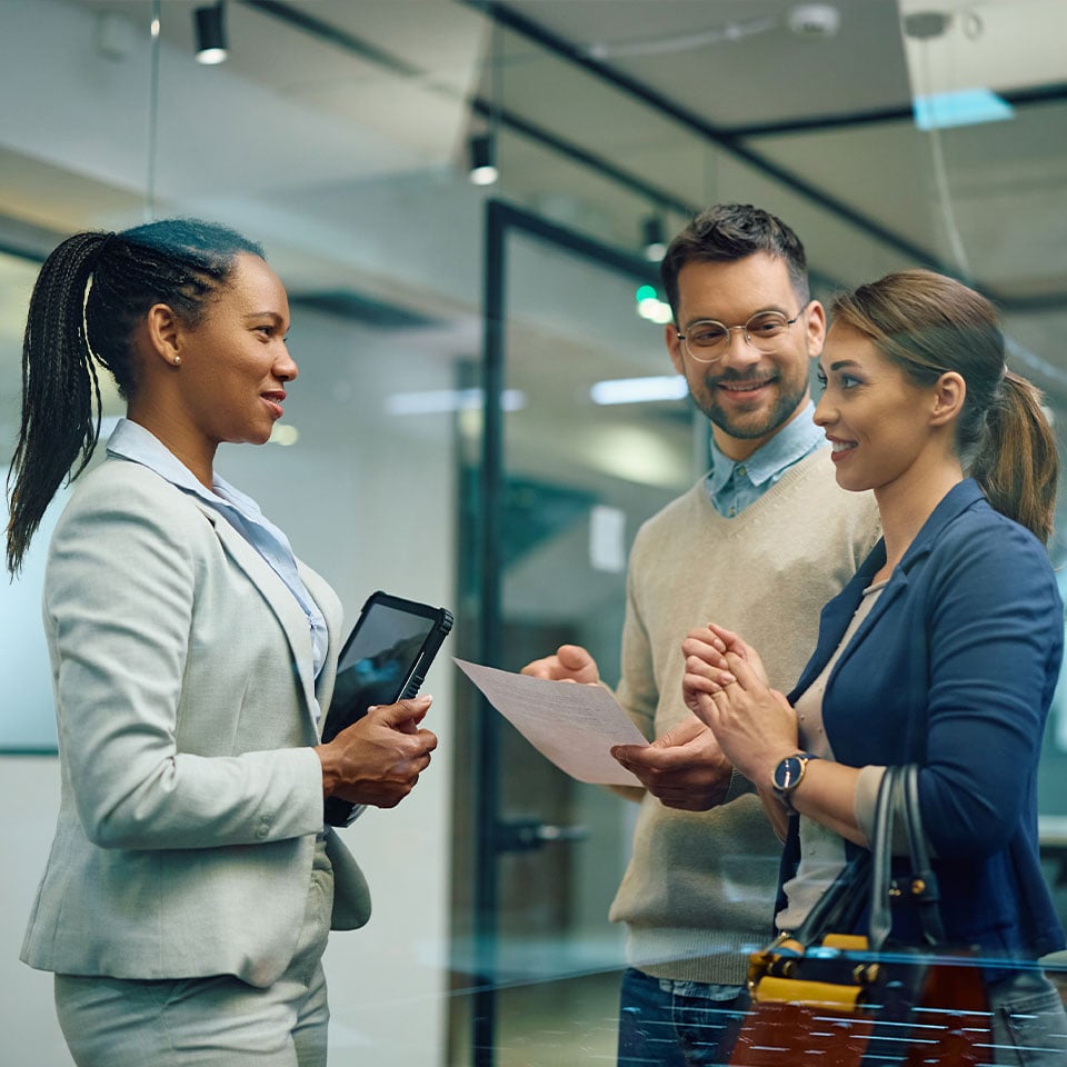 Three people in business attire are having a discussion in an office setting. One person is holding a tablet, another is holding a piece of paper, and the third person has a handbag. The image highlights IT services for financial industry